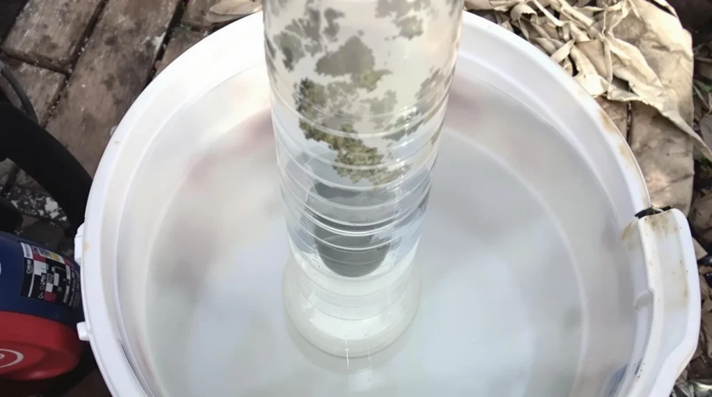 Homemade gravity bong setup in use, showing a plastic bottle in a bucket filled with water, with visible smoke inside the bottle.