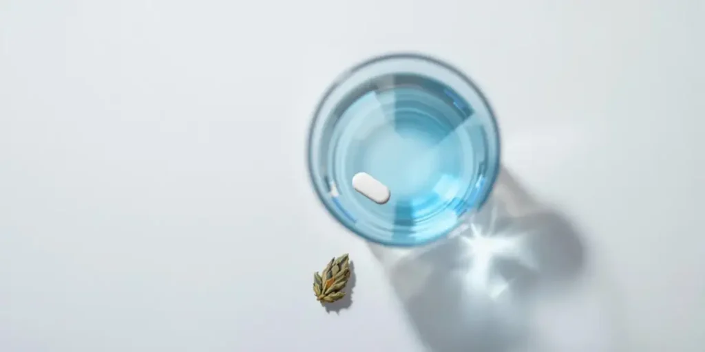 glass of water with pill and cannabis bud on white background