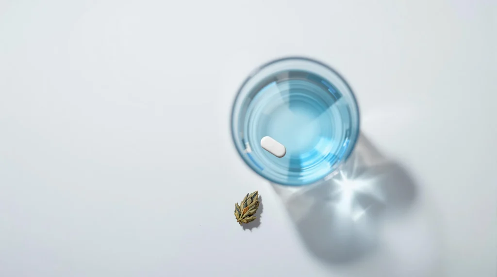 CBD oil bottle and ibuprofen pill next to a glass of water on a white surface.