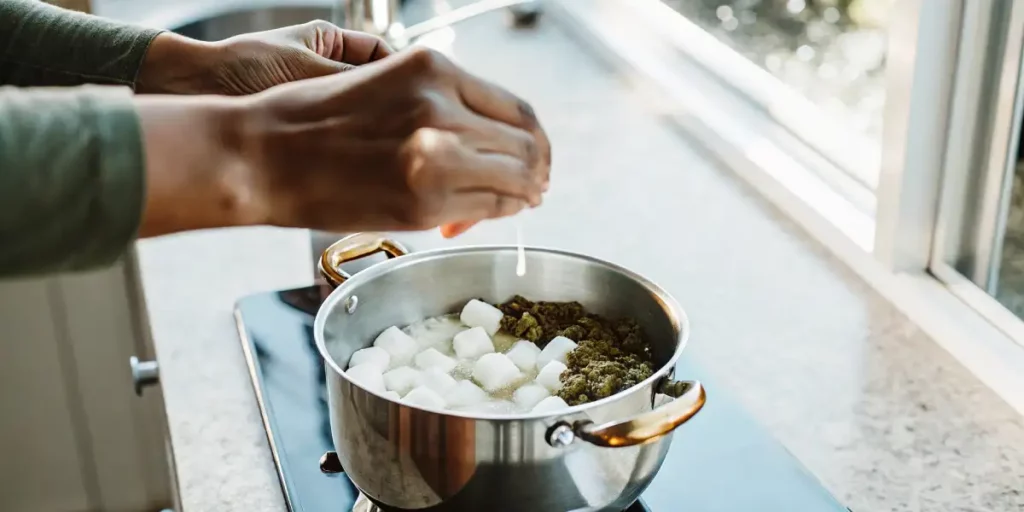 Hands gently mixing marshmallows and ground cannabis in a pot for homemade edibles