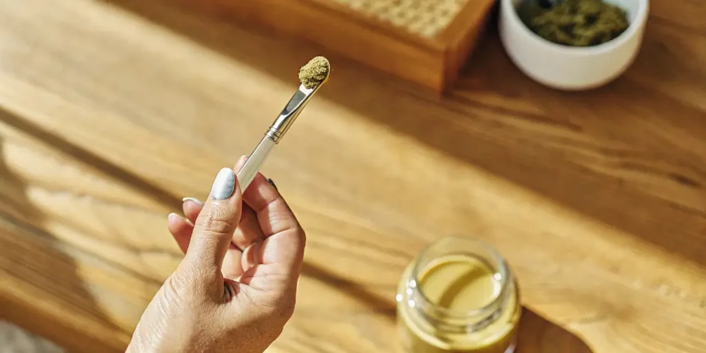 Hand holding dab tool with kief-covered concentrate above a wooden table with jars and grinder.