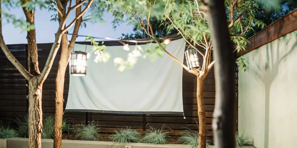 Outdoor garden with white projector screen hanging between trees, surrounded by string lights and wooden fence.