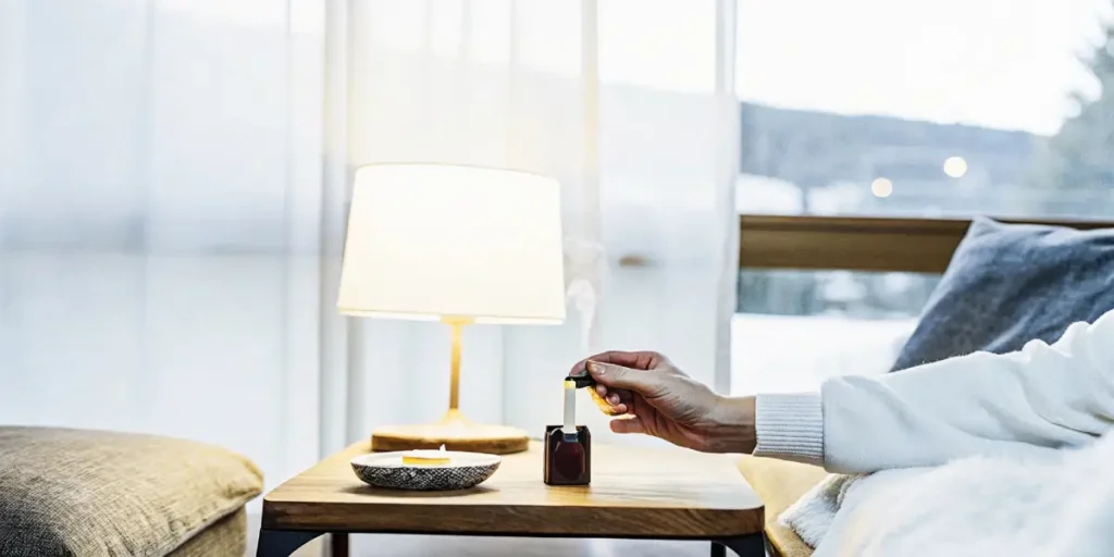 Person's hand using an aromatherapy stick near a lit lamp on a wooden side table, in a cozy room at dusk.