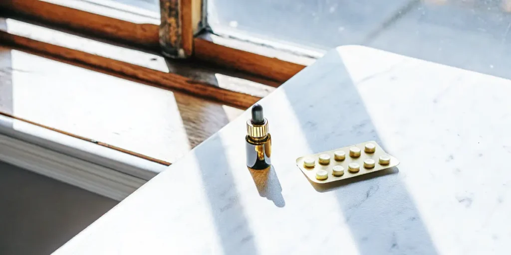 Close-up of a brown CBD oil dropper bottle and a blister pack of pills on a white marble tabletop, illuminated by strong sunlight.