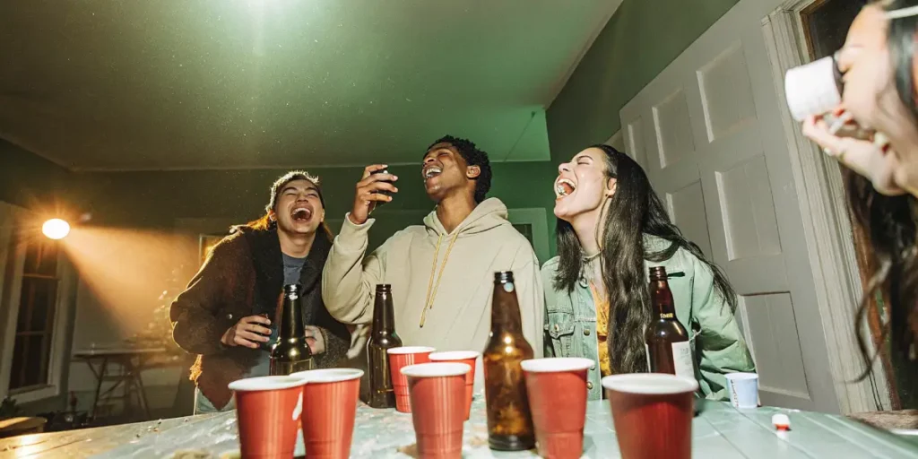 Group of friends laughing loudly around a table with red cups and beer bottles, under green party lights.