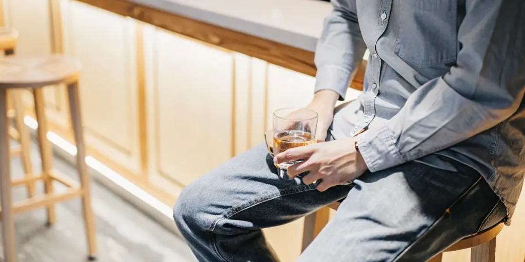 Young man sitting on a bar stool, holding a glass with an amber drink, in a brightly lit bar setting.