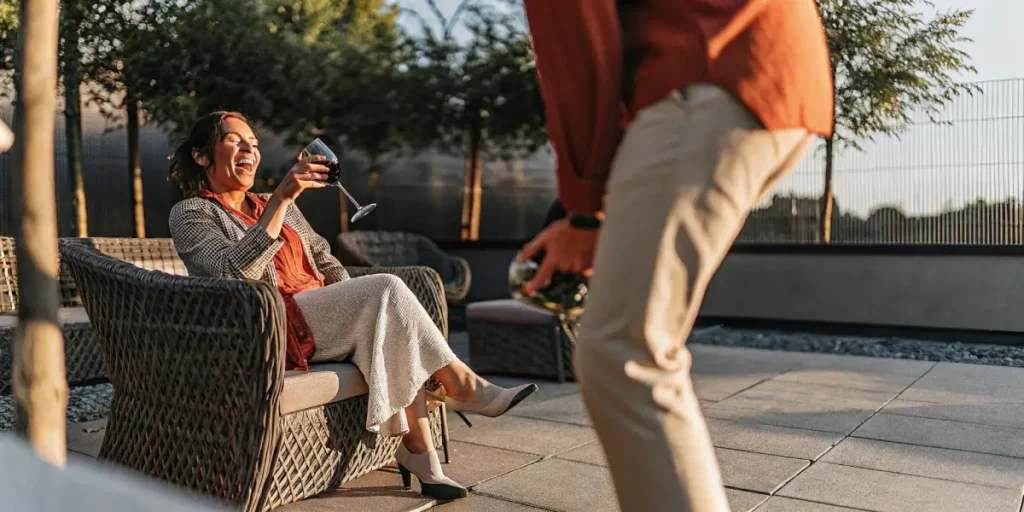 Woman laughing, raising wine glass in a wicker chair at an outdoor garden party, with another person and trees in background.