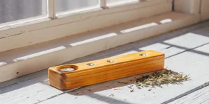 Close-up of a handcrafted wooden 3-hole hash pipe on a wooden windowsill with dried herbs and sunbeams.