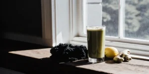 Glass of green detox juice with kale, lemon, and ginger on a wooden windowsill.